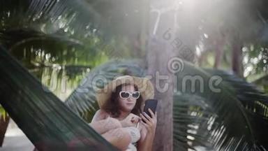 戴着太阳镜和帽子的放松的女孩用手机躺在棕榈树之间的吊床上。 <strong>1920</strong>x1080